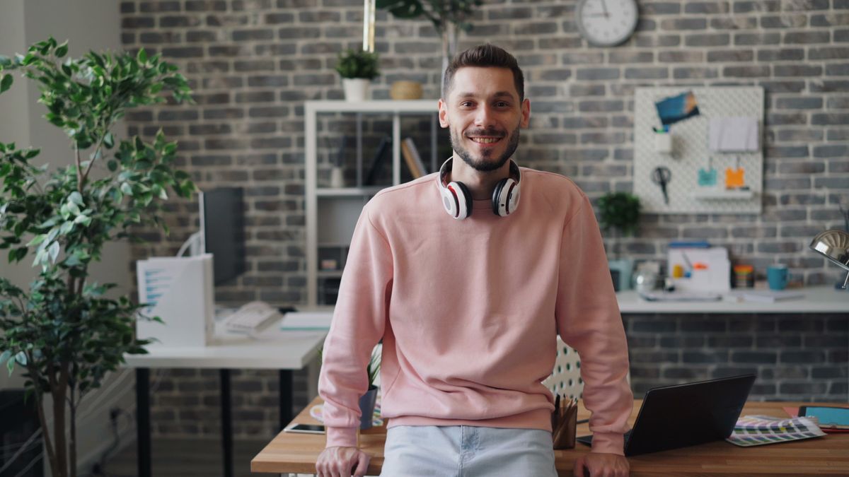 A solo entrepreneur smiling in his home studio, laptop open on the desk behind him